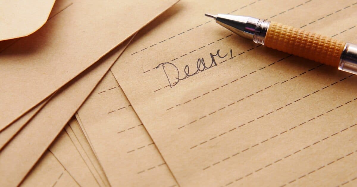 A pen resting on lined notepaper reading "Dear", on a stack of kraft envelopes